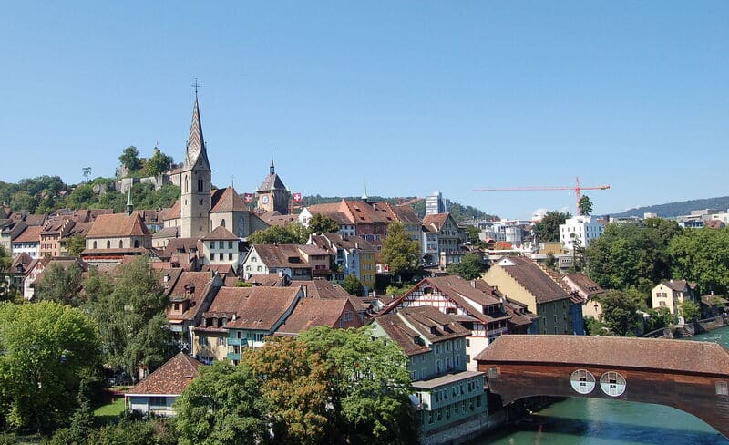 Altstadt im Aargau mit Kirche, dicht stehenden Häusern und überdachter Brücke am Fluss.