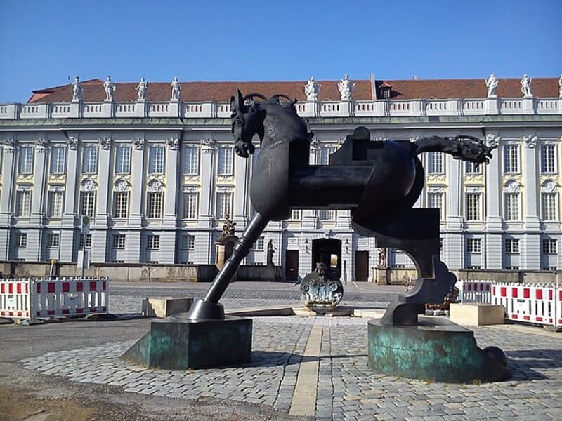 Dunkle Metallskulptur eines Pferdes vor dem verzierten Schlossgebäude von Ansbach bei Tageslicht.