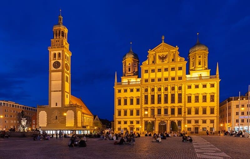 Coreflow-Augsburg-Hero Abendliche Ansicht des beleuchteten Rathauses von Augsburg mit Perlachturm und Menschen auf dem Platz.