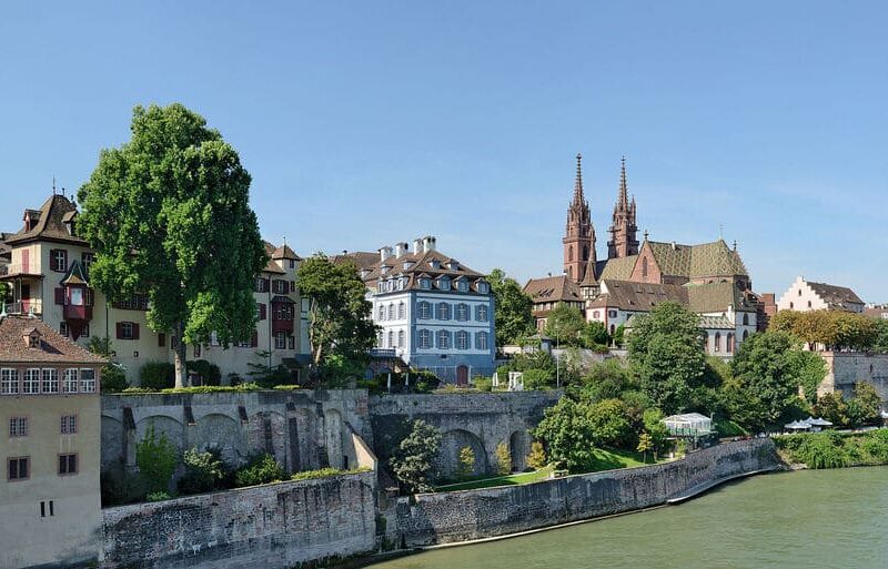 Uferansicht von Basel mit historischen Gebäuden, viel Grün und dem Münster über der Rheinkante.
