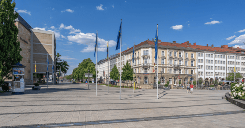 Weitläufiger Platz in Bayreuth mit Flaggenmasten, historischen Fassaden und blauem Himmel.
