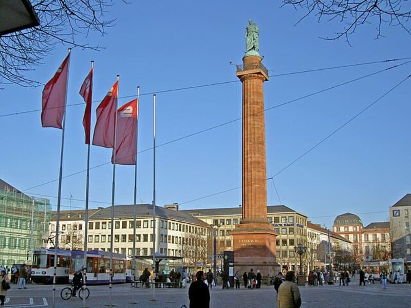 Hohe Säule mit Statue auf dem Luisenplatz in Darmstadt, umgeben von Stadtgebäuden und Flaggen.