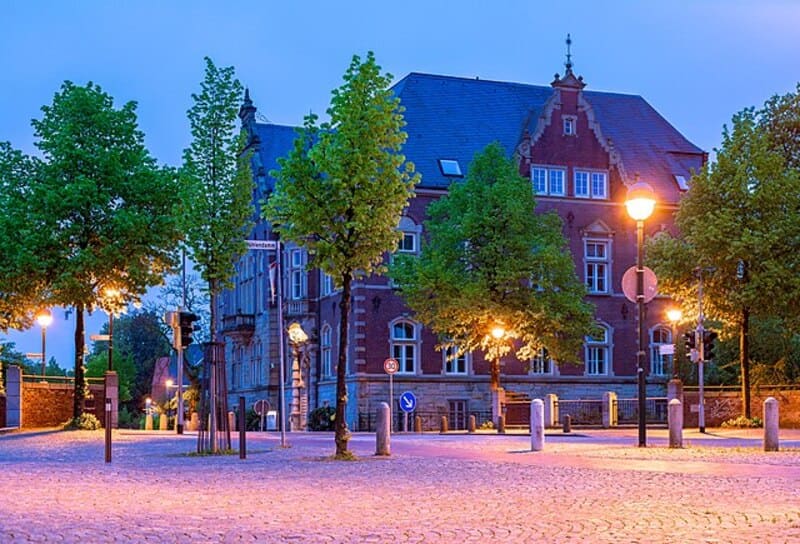 Platzansicht in Delmenhorst bei Dämmerung mit Kopfsteinpflaster, altem Gebäude und warmem Straßenlicht.