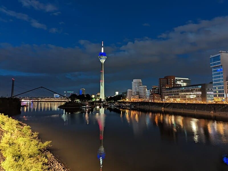 Nächtliche Skyline von Düsseldorf mit beleuchtetem Rheinturm und Reflexionen im Wasser.
