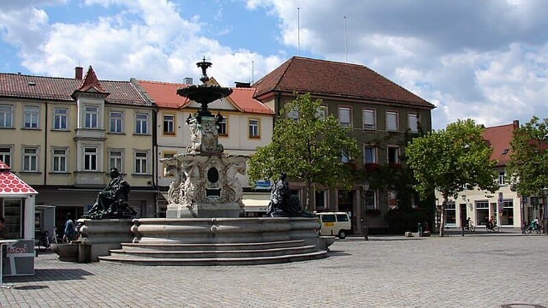 Coreflow-Erlangen-Hero Marktplatz in Erlangen mit historischem Brunnen, umliegenden Gebäuden und Bäumen bei Sonnenschein.