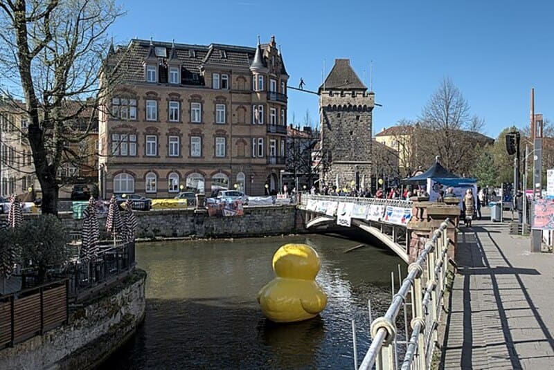 Flussufer in Esslingen mit historischer Brücke, Turm und großer gelber Gummiente im Wasser.