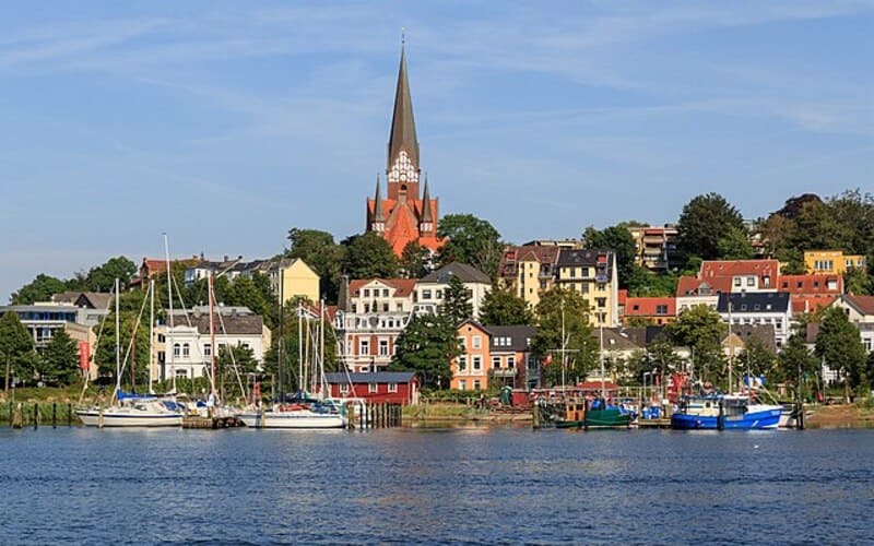 Blick auf Flensburgs Hafen mit Segelbooten und farbigen Altstadthäusern vor Kirchturm.
