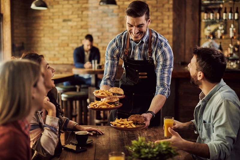 Servicekraft bringt frisch zubereitete Burger zu Gruppe in gemütlicher Bar mit Backsteinwand.