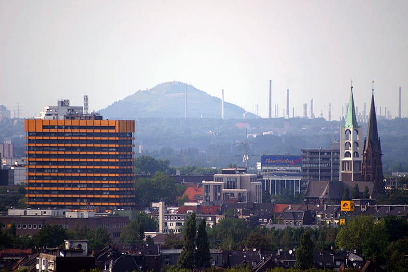 Stadtansicht von Gelsenkirchen mit markantem orangefarbenem Hochhaus und Kirchtürmen im Vordergrund.