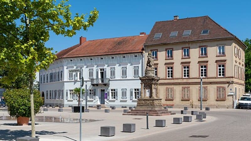 Denkmal auf offenem Platz vor hellen Verwaltungsgebäuden und Bäumen in Germersheim.