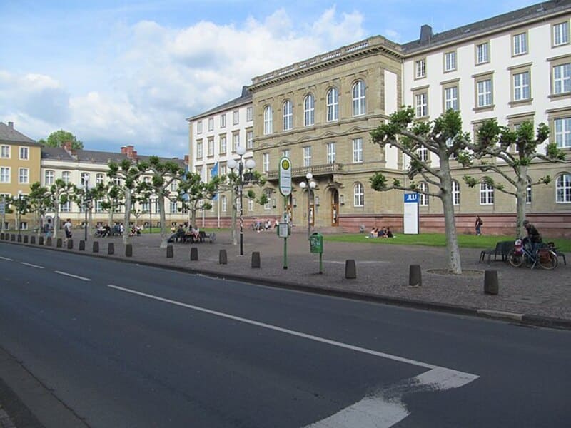 Historischer Gebäudekomplex der Universität Gießen mit Platz, Bäumen und wartenden Personen an der Haltestelle.