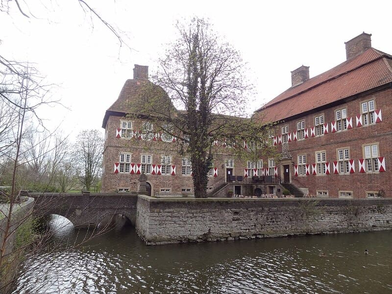 Coreflow-Hamm-Hero Mehrstöckiger Bau mit roten Fensterläden und zentralem Baum am Wassergraben in Hamm.