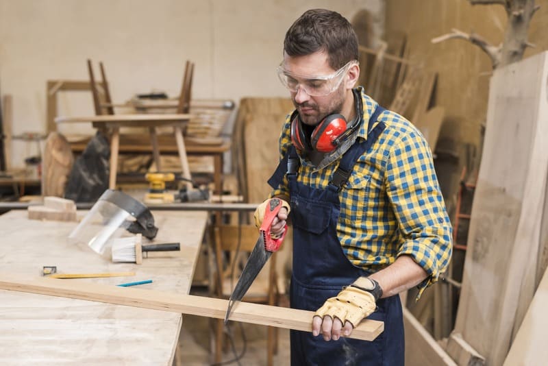 Coreflow-Handwerker-Hero Handwerker bearbeitet Holz mit Handsäge, trägt Gehörschutz und Arbeitshandschuhe im Innenraum.
