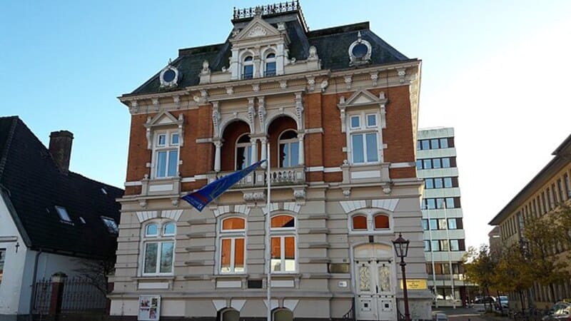 Historisches Verwaltungsgebäude in Heide mit verzierten Fassadenelementen und wehender Flagge im Vordergrund.