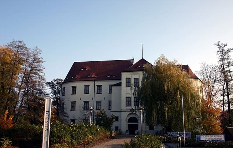 Historisches Schloss Hoyerswerda mit hellem Fassadenbau, rotem Dach und umgebenden Bäumen im Herbstlicht.