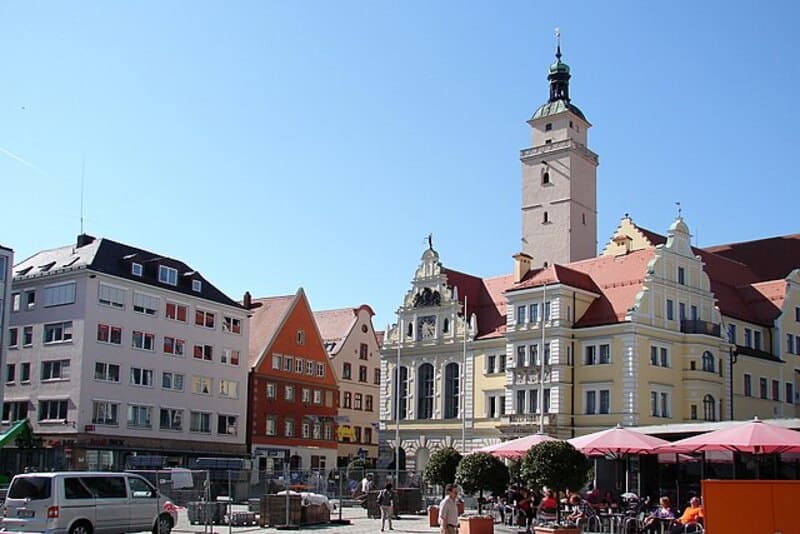 Historische Gebäudereihe mit farbigen Fassaden und hohem Turm auf einem sonnigen Platz in Ingolstadt.