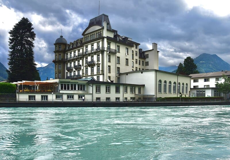 Großes helles Hotelgebäude am Wasser in Interlaken, Schweiz, vor bergiger Landschaft.