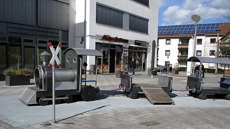 Coreflow-Iserlohn-Hero Spielbereich mit kleiner Metalllok und Waggons auf einem Platz vor Geschäften in Iserlohn.