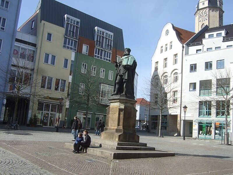 Statue auf dem Marktplatz von Jena, umgeben von modernen und historischen Gebäuden.