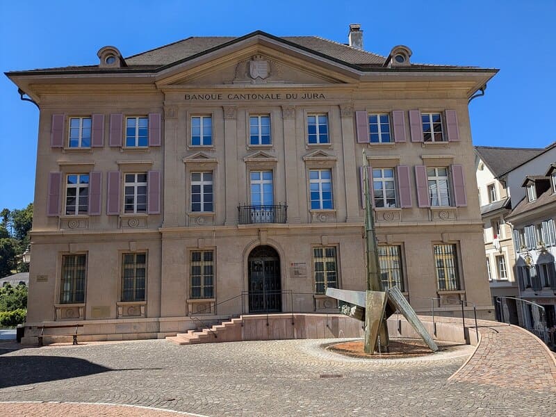 Historische Fassade der Banque Cantonale du Jura mit Brunnenplastik auf kleinem Platz.
