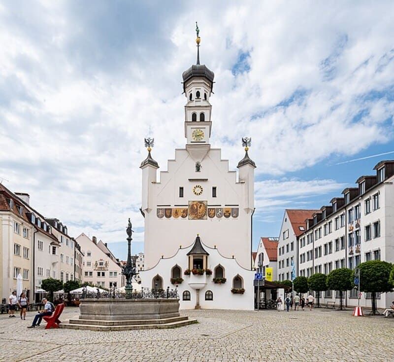Markantes Rathaus mit Uhr, Figurenfenstern und Brunnen im Vordergrund im Zentrum von Kempten.