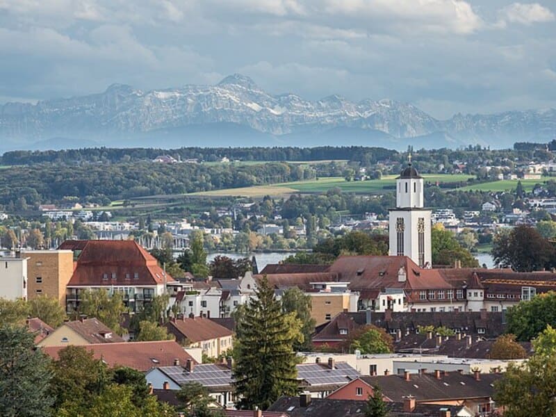 Blick über die Dächer von Konstanz mit weißem Kirchturm und Alpenkulisse im Hintergrund.