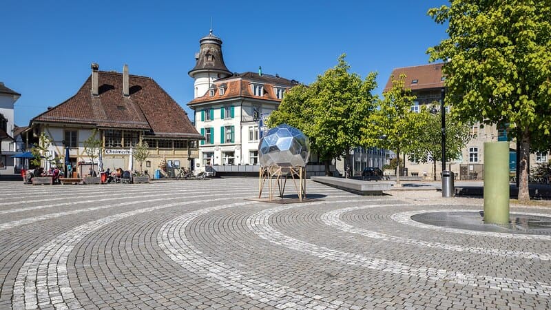 Coreflow-Langenthal-Hero Offener Platz in Langenthal mit Brunnenobjekt, Bäumen und umgebenden Altstadtbauten.