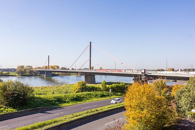 Schrägseilbrücke über den Rhein mit herbstlicher Uferlandschaft und vorbeifahrenden Autos in Leverkusen.