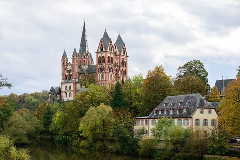 Blick auf den Limburger Dom über herbstlichem Baum­bestand und Gebäuden am Flussufer.