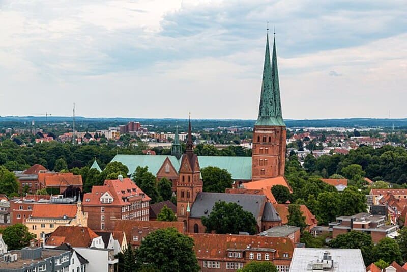 Blick über Lübecks Altstadt mit hohen Kirchtürmen und weitläufiger Umgebung unter wolkigem Himmel.