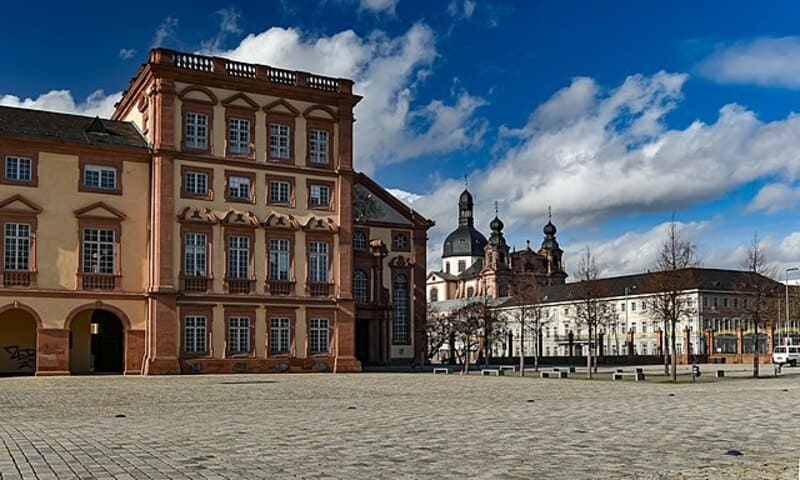 Historisches Mannheimer Gebäude mit roter Sandsteinfassade vor großem, leerem Platz und blauem Himmel.