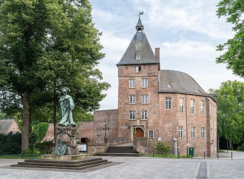 Historisches Backsteingebäude mit Turm und davorstehender Statue auf offenem Platz in Moers.