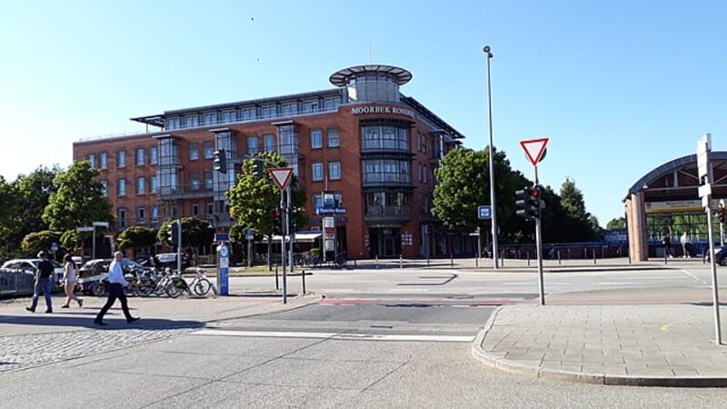 Modernes Eckgebäude in Norderstedt an Kreuzung mit Verkehrsschildern, Bäumen und Passanten.