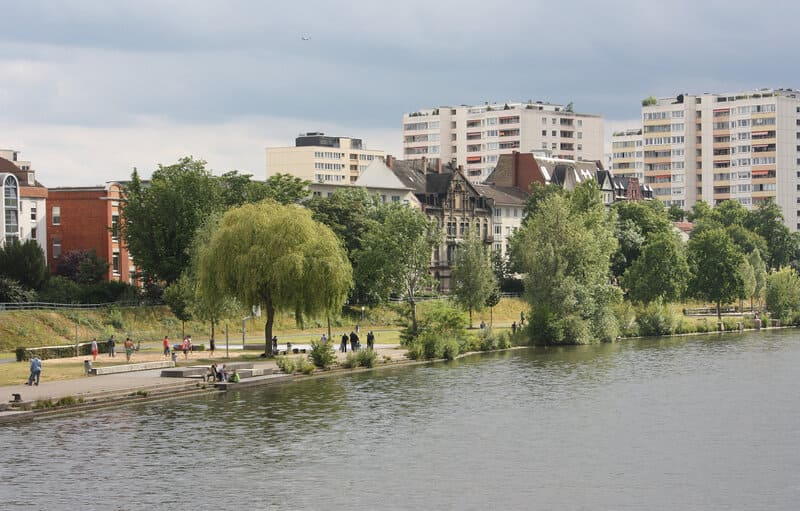 Flussufer in Offenbach mit Spaziergängern, Bäumen und Wohnhochhäusern im Hintergrund.