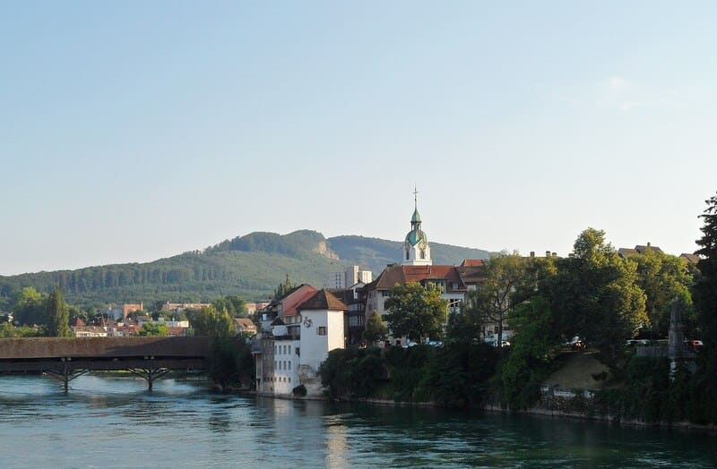 Blick auf Olten mit Aare, gedeckter Brücke, Altstadthäusern und bewaldetem Hügel.
