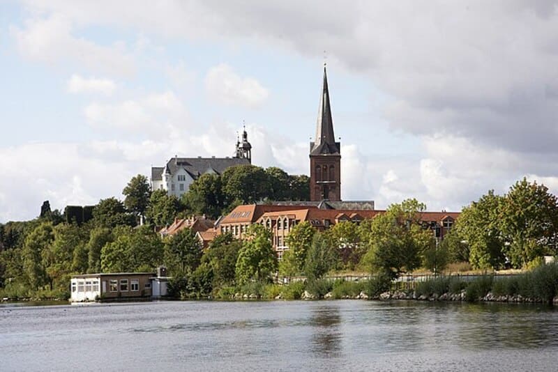 Uferlandschaft in Plön mit Wasserfläche, grünen Bäumen und markantem Kirchturm im Hintergrund.