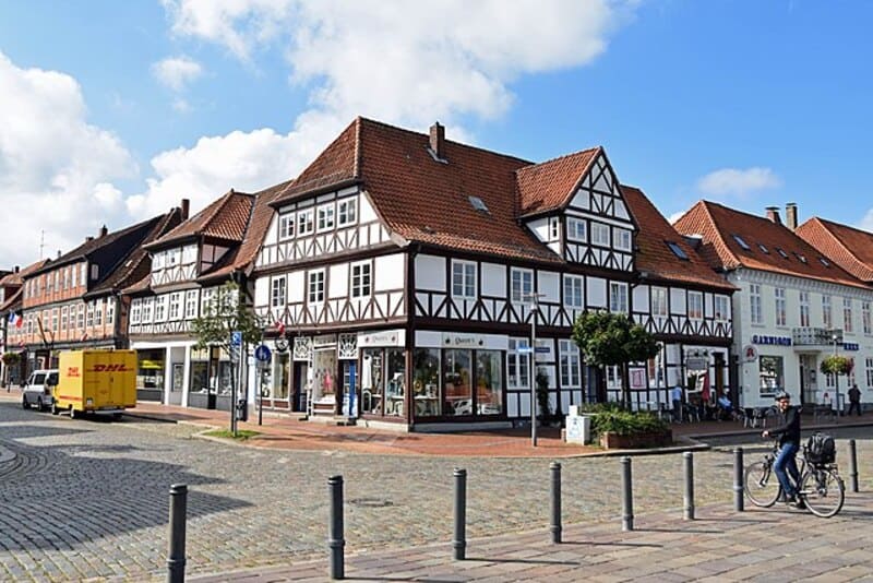 Fachwerkhaus in Rendsburg an gepflastertem Platz mit Radfahrer und gelbem Lieferwagen im Sonnenlicht.