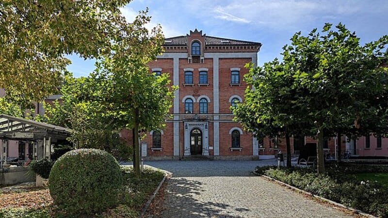 Historisches Backsteingebäude in Rosenheim, flankiert von Bäumen und gepflastertem Weg im Vordergrund.