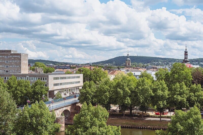 Blick über Saarbrücken mit Brücke, dichtem Baumbestand und Stadtansicht vor hügeliger Landschaft