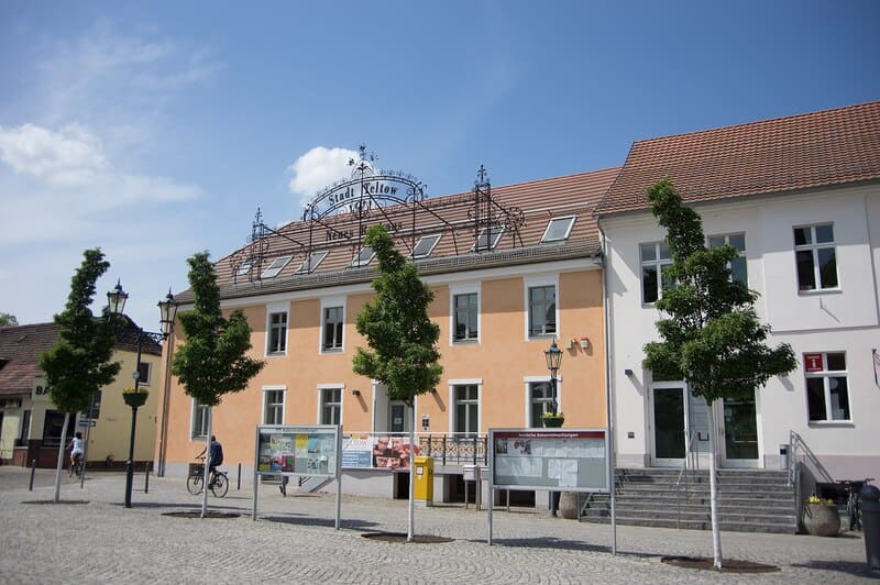 Helles Rathaus Teltow mit Vorplatz, Bäumen und Infotafeln an einem sonnigen Tag.