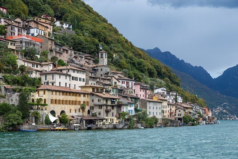 Uferdorf im Tessin mit dicht stehenden Häusern am See und bewaldetem Berghang.