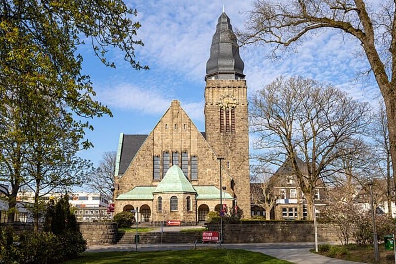 Historische Sandsteinkirche in Velbert bei Tageslicht, umrahmt von kahlen und grünen Bäumen.
