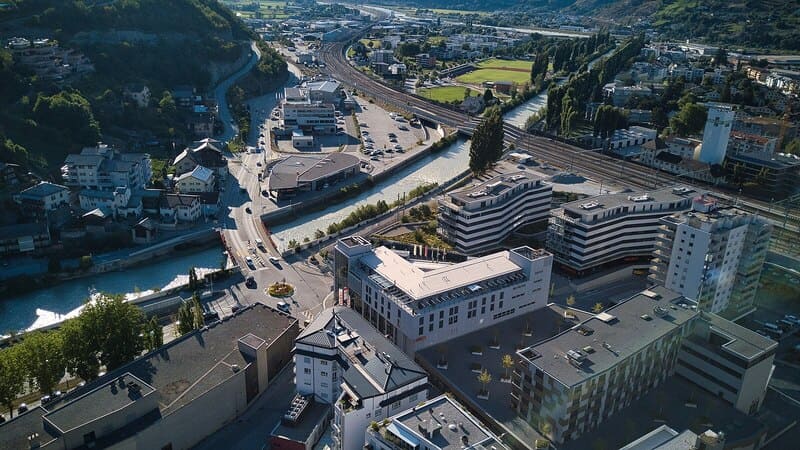 Luftaufnahme von Visp, Schweiz, mit modernen Gebäuden, Flusslauf, Straßen und Bahntrassen im Tal.