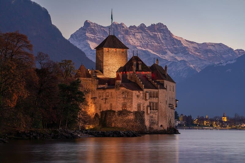 Beleuchtete Burg im Kanton Waadt am Seeufer mit Alpen im Hintergrund bei Abenddämmerung.