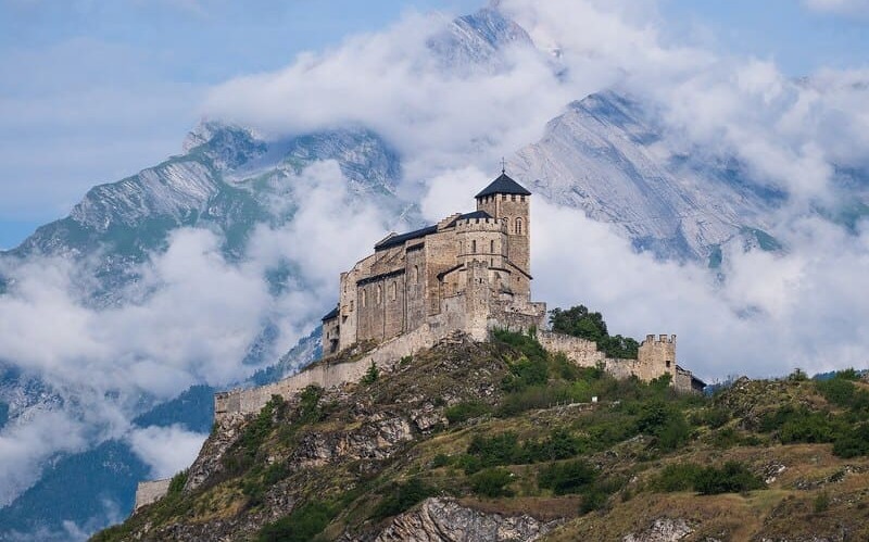 Historische Burg im Wallis über dem Tal, umgeben von Bergen und aufziehendem Nebel.