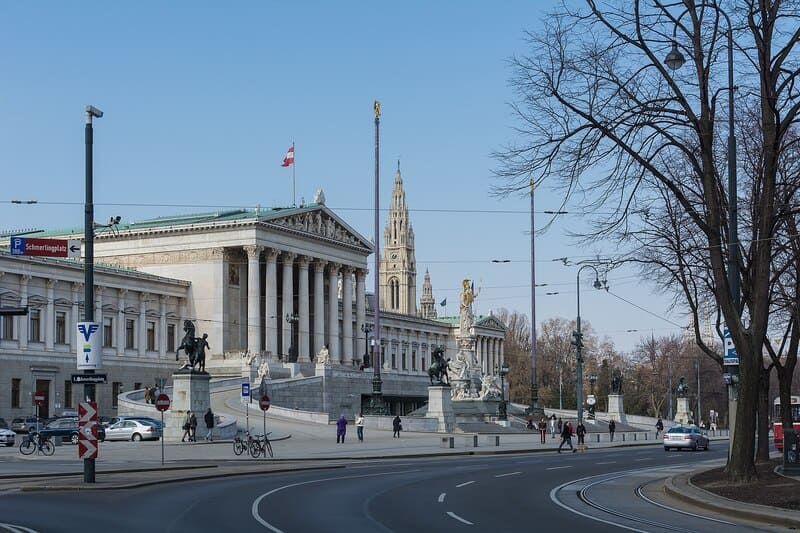 Foto des österreichischen Parlamentsgebäudes in Wien mit Säulenfassade, Statuen und Straßenverkehr im Vordergrund.