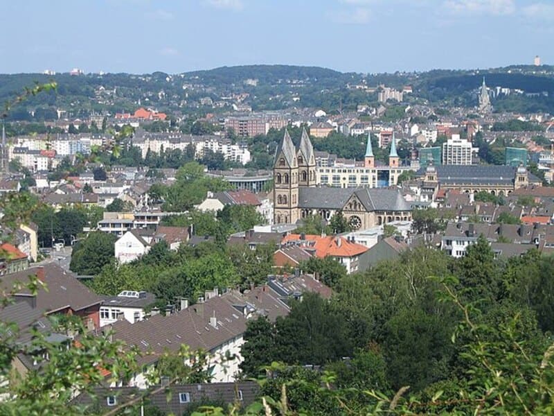 Panorama von Wuppertal mit dicht bebauten Wohngebieten, Kirche und bewaldeten Hügeln im Hintergrund.