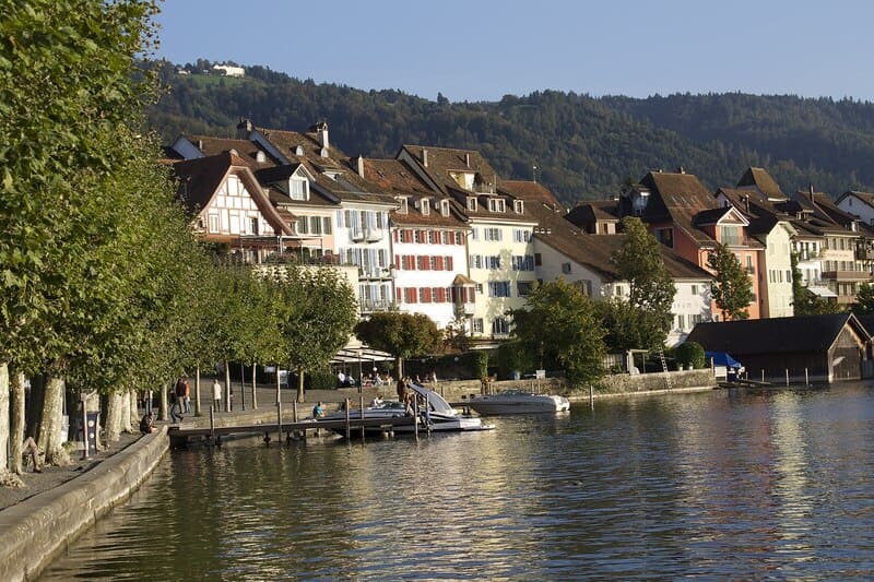 Altstadt von Zug mit farbigen Häusern am Seeufer, Bootssteg und ruhigem Wasser.