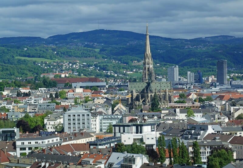 Blick über Linz mit markantem Kirchturm, Wohngebieten und grüner Hügellandschaft unter bewölktem Himmel.