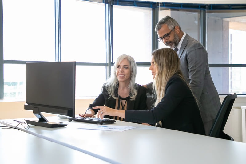Drei Personen besprechen gemeinsam Inhalte am Computerarbeitsplatz in modernem Büro mit großen Fenstern.